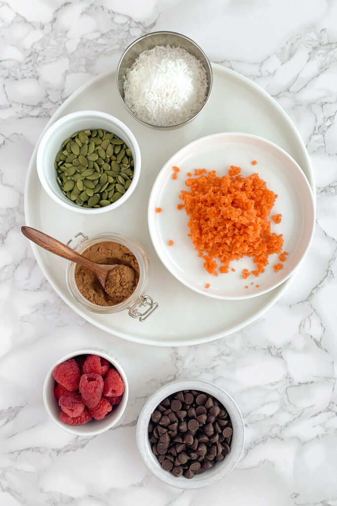 examples of different add-in ingredients in bowls; shredded carrots, raspberries, chocolate chips, shredded coconut and pumpkin seeds. 