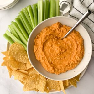 dairy free buffalo chicken dip in a bowl with spoon, surrounded by celery and tortilla chips.