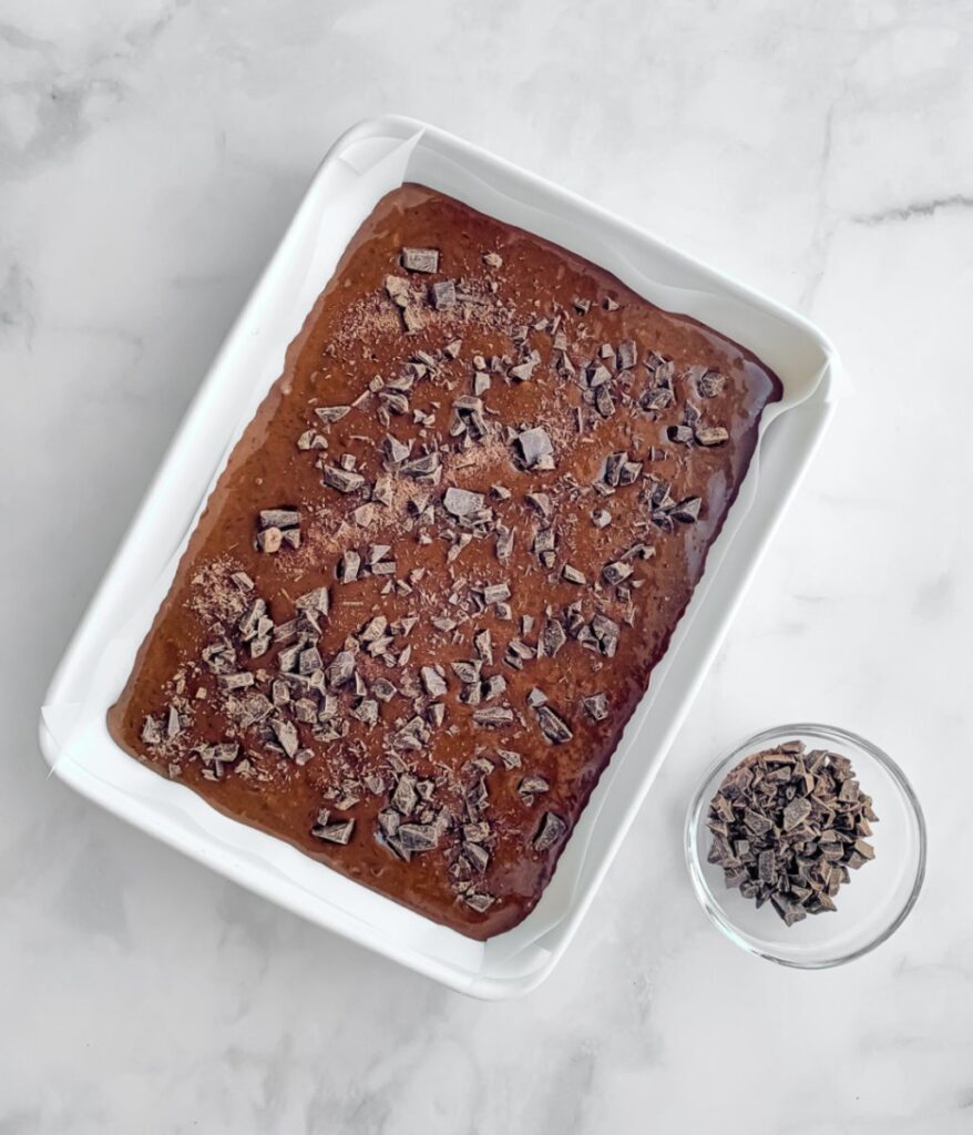 brownie batter in a white ceramic dish, sprinkled with chopped chocolate and chopped chocolate in a separate bowl.