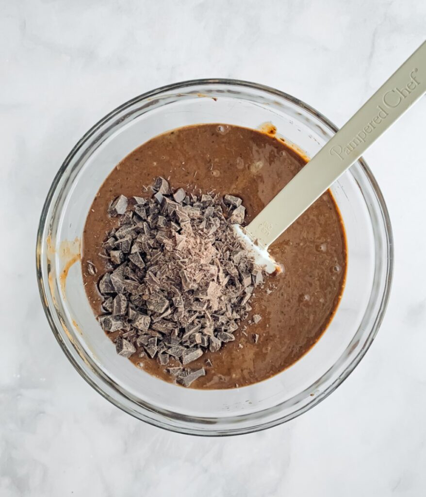 brownie batter in a bowl with chopped chocolate on top and spatula, ready to mix.