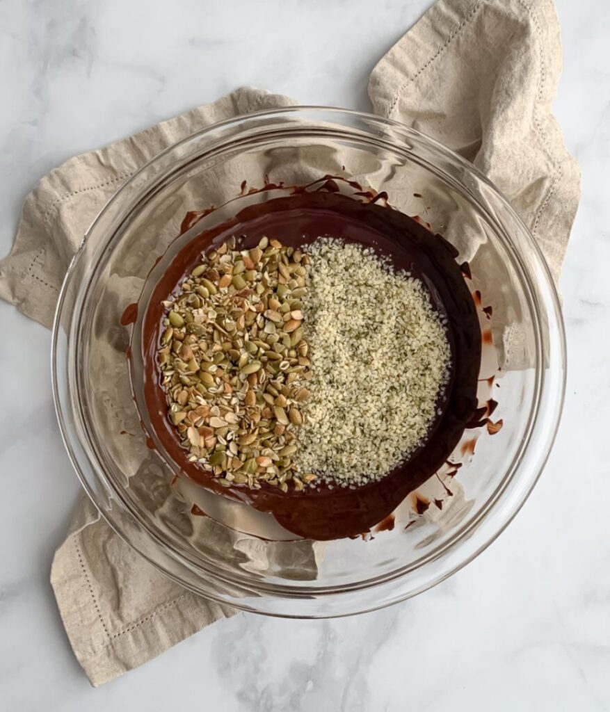 melted chocolate in a glass bowl on a tea towel, with seeds on top about to be stirred in.