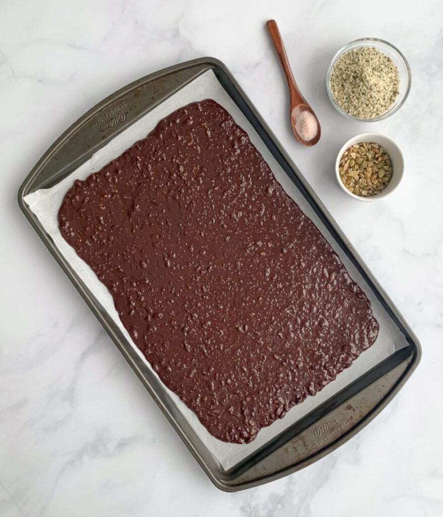 melted chocolate with seeds on a parchment lined pan ready to be topped with more seeds. pumpkin seeds, hemp hearts and pink salt in glass bowls beside the pan.