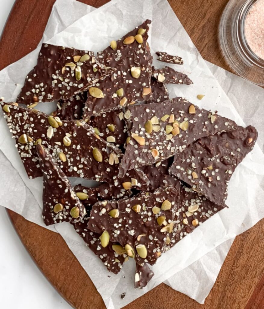 pile of broken up magnesium chocolate bark with seeds on top, on top of parchment paper on a cutting board.