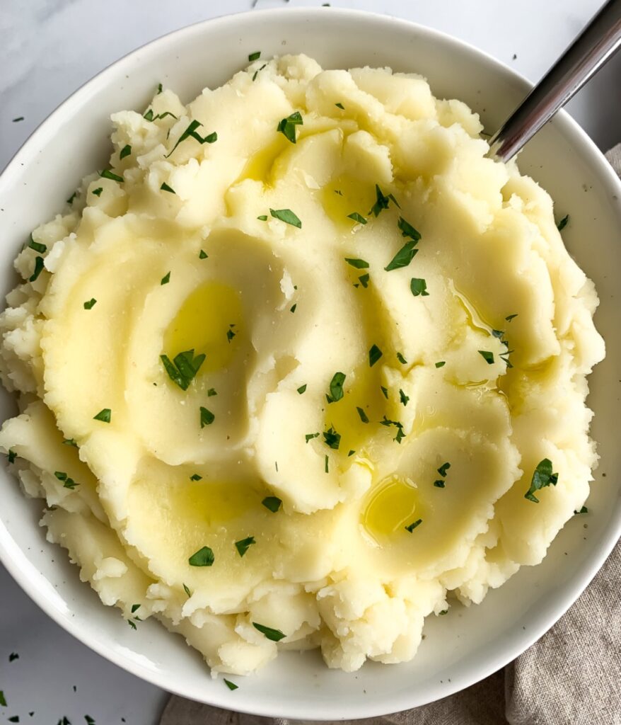mashed potatoes in an off-white bowl with a serving spoon, garnished with olive oil and chopped parsley