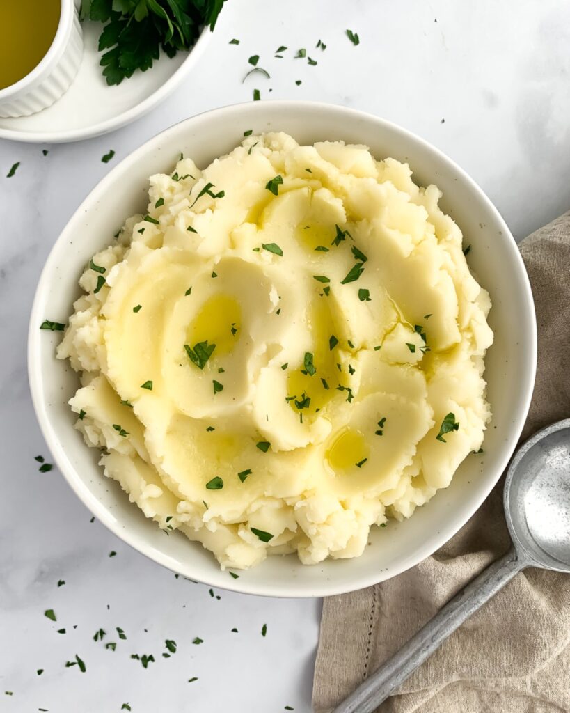 mashed potatoes in an off-white bowl garnished with olive oil and chopped parsley, with olive oil and leafy parsley in the top left corner and cloth and silver spoon in the bottom right