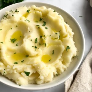 mashed potatoes in an off-white bowl garnished with olive oil and chopped parsley, with olive oil and leafy parsley
