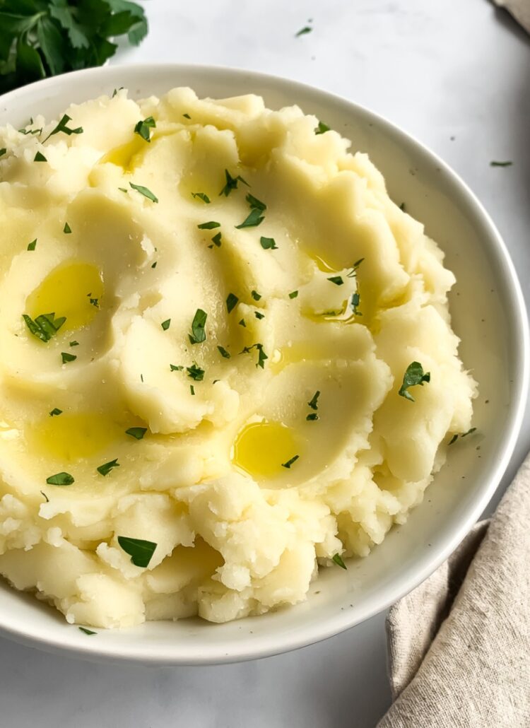 mashed potatoes in an off-white bowl garnished with olive oil and chopped parsley, with olive oil and leafy parsley