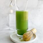 matcha soda in tall ribbed glass on small white plate with matcha bamboo whisk and bottle of water in background.