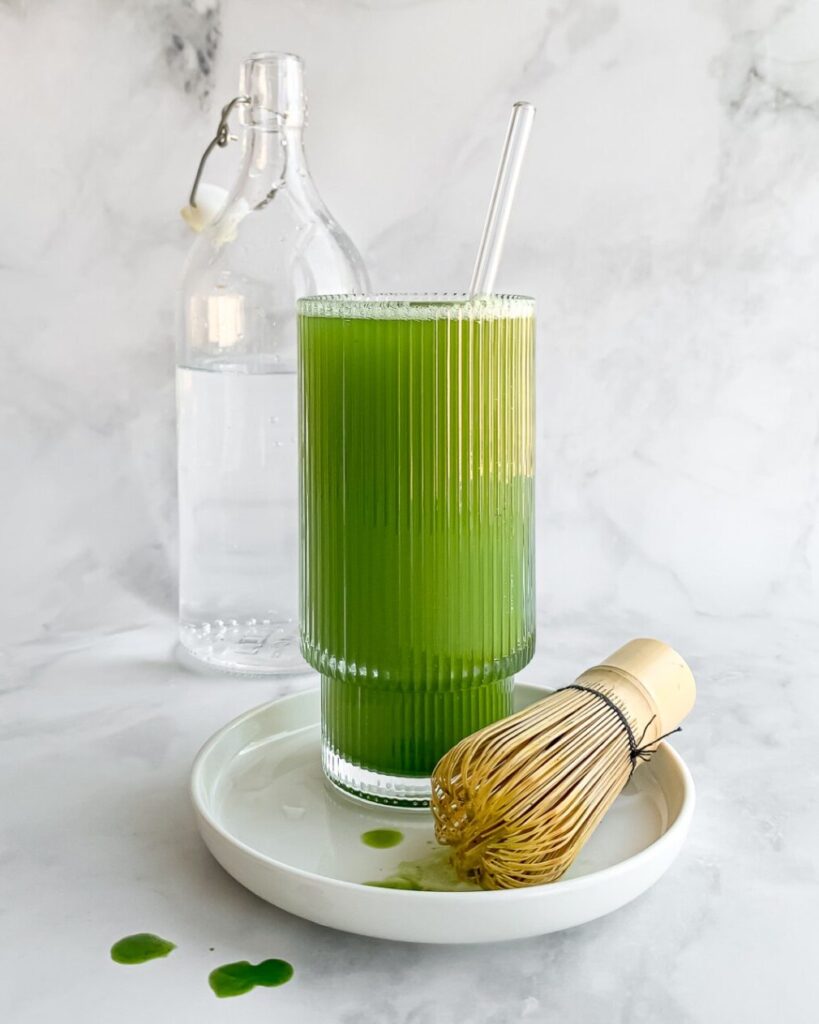 matcha soda in tall ribbed glass on small white plate with matcha bamboo whisk and bottle of water in background.