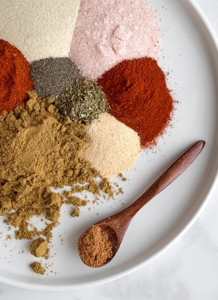 eight different spices on a white plate and a wooden spoon holding gluten free taco seasoning
