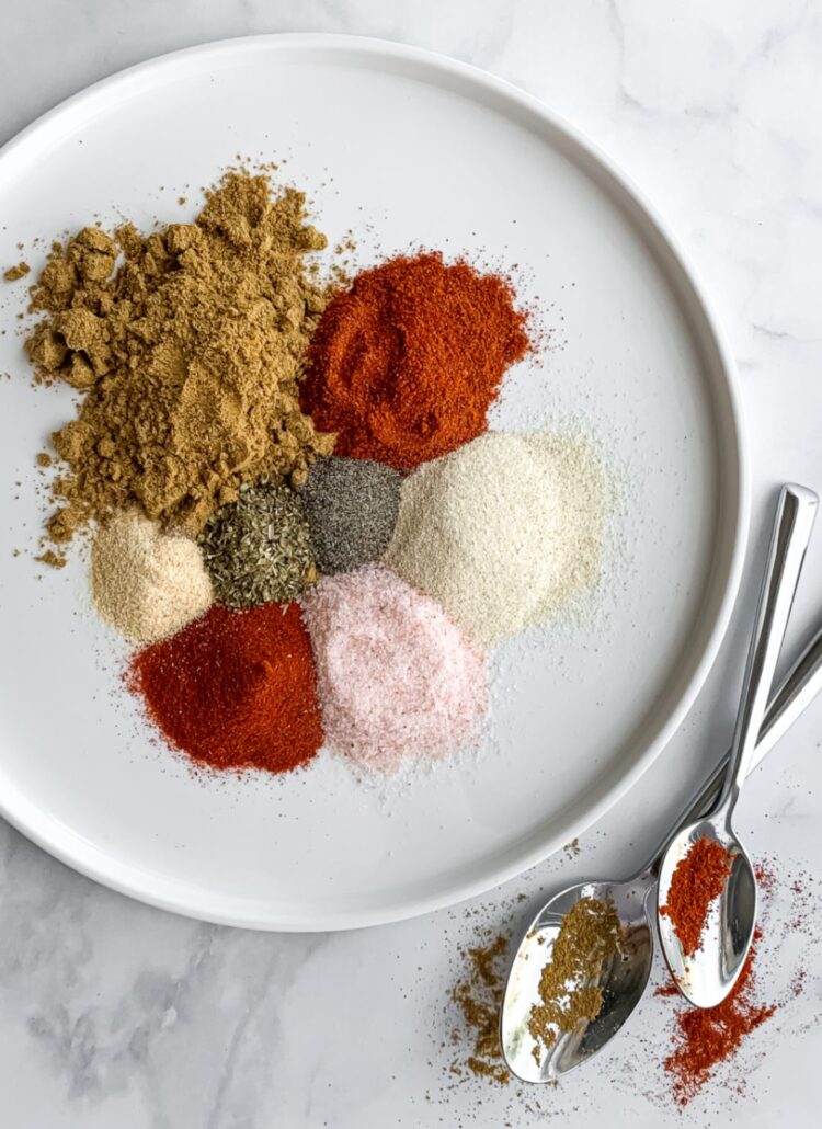 eight different spices on a white plate with two spoons beside it
