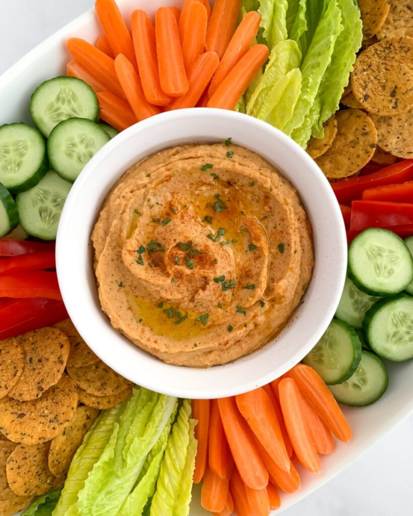 taco hummus garnished with cilantro and olive oil, in a white bowl surrounded by vegetables on platter