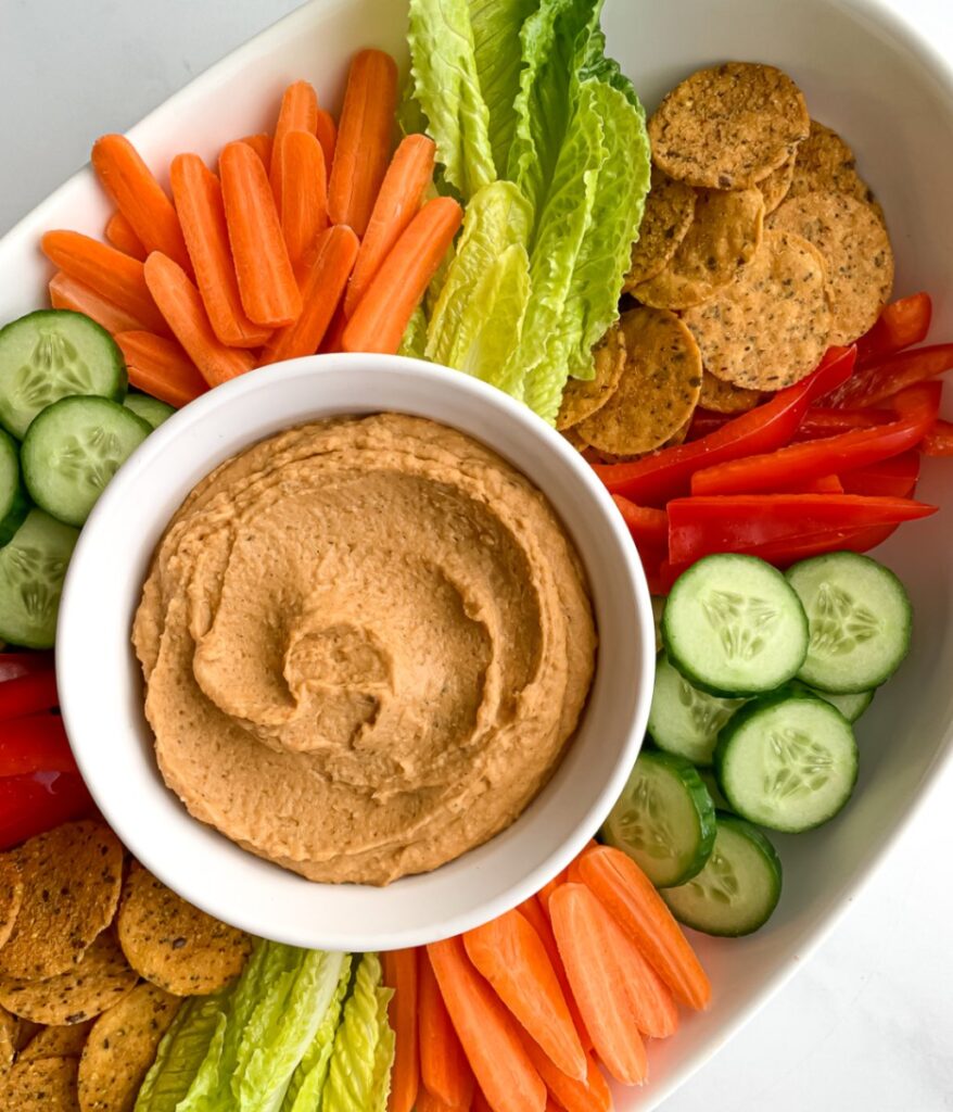 creamy taco hummus in a white bowl surrounded by vegetables and crackers on a white platter