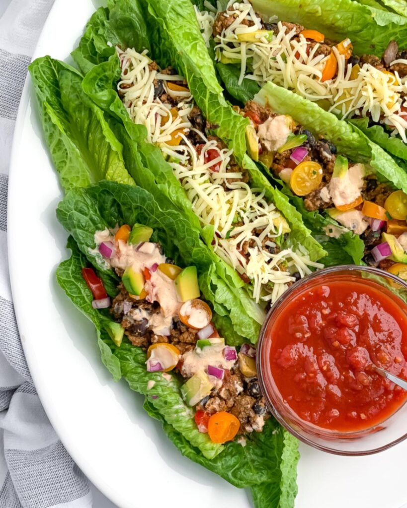 4 rainbow taco salad boats on a white plate with a small bowl of salsa