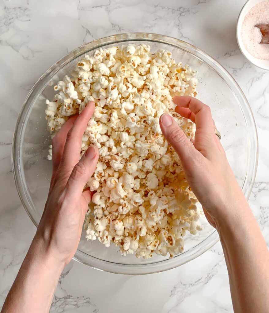 hands tossing popcorn in a glass bowl