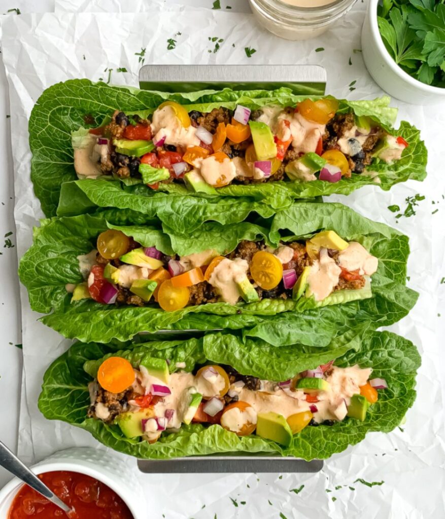 3 rainbow taco lettuce boats in taco holder with salsa, dairy free cheese sauce and cilantro in small bowls