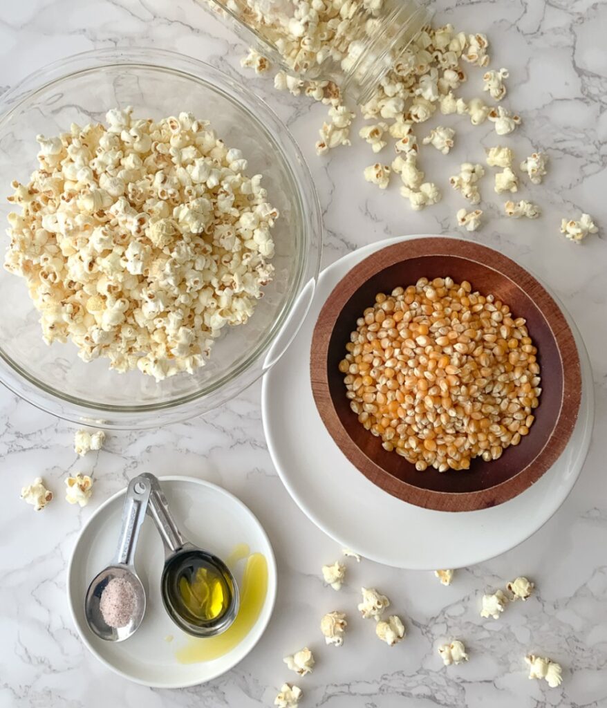 popcorn kernels in a wooden bowl on a white plate, spoons on a white plate, each with olive oil and salt and popcorn in a glass bowl