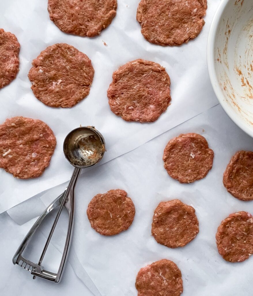 raw chicken sausage patties on parchment paper with a white bowl and cookie scoop