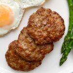 white plate with 2 fried eggs 3 chicken sausage patties and steamed asparagus