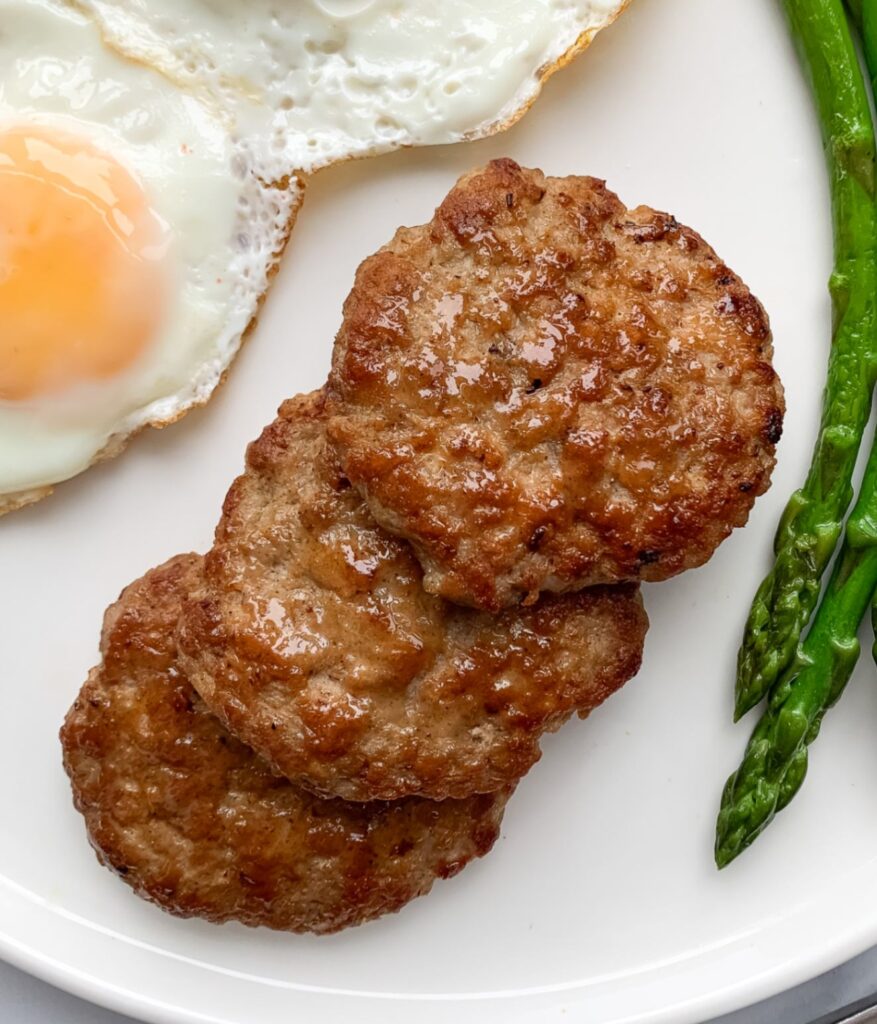 white plate with 2 fried eggs 3 chicken sausage patties and steamed asparagus