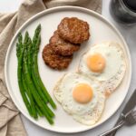 white plate with 2 fried eggs 3 chicken sausage patties and steamed asparagus and black coffee to the side