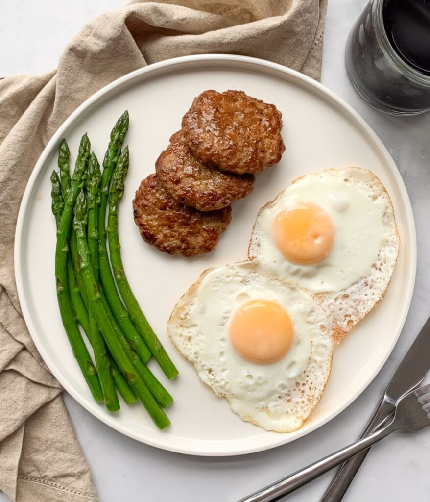 white plate with 2 fried eggs 3 chicken sausage patties and steamed asparagus and black coffee to the side