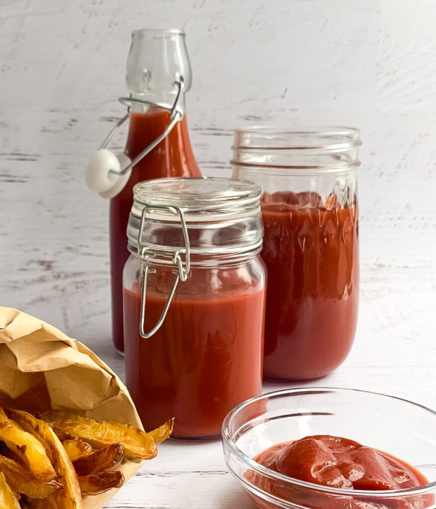 3 different looking glass jars with ketchup , small bowl of ketchup and french fries in a brown paper bag