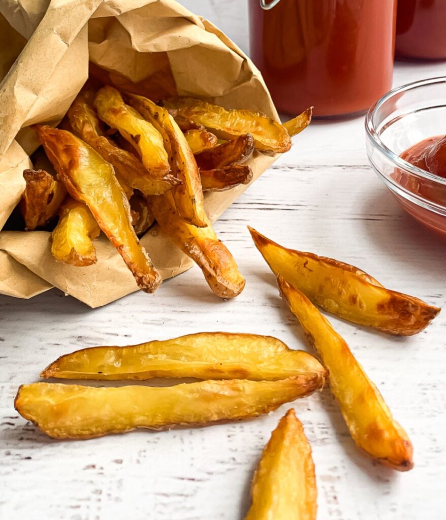 crispy potato wedges falling out of a small brown paper bag, with glass jars and a bowl with ketchup to the side