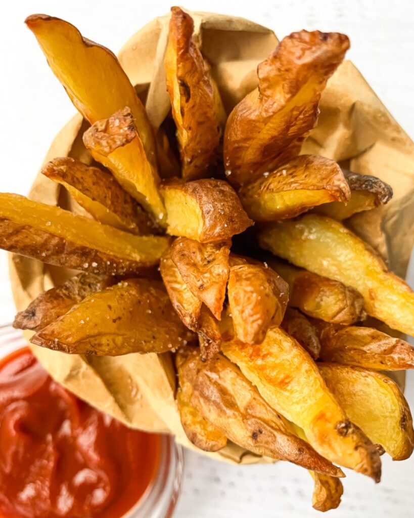 top down shot of potato wedge fries in a small brown paper bag with ketchup in a small bowl beside it