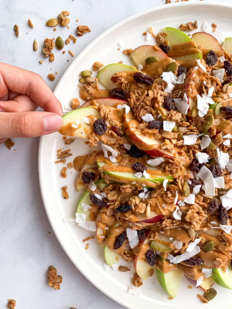 Hand reaching for apple nachos topped with peanut butter, granola, seeds, and raisins.