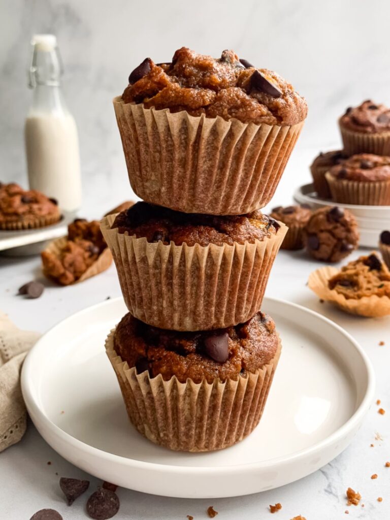 3 chocolate chip gluten-free banana zucchini muffins made with almond flour stacked on top of each other on a plate with a bottle of milk and other muffins in the background.