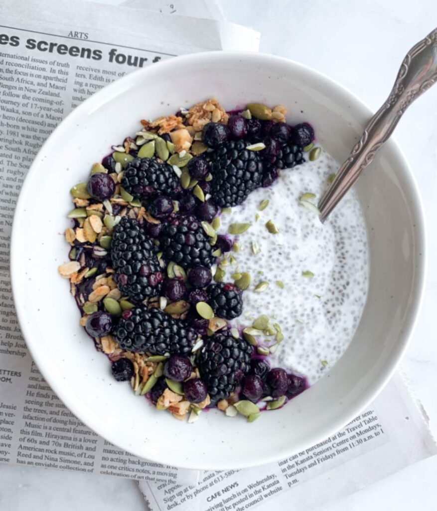 prepared chia pudding bowl topped with granola, pumpkins seeds, fresh blackberries and frozen blueberries in a crescent moon patterns, with a spoon, placed on top of a folded page of newspaper.