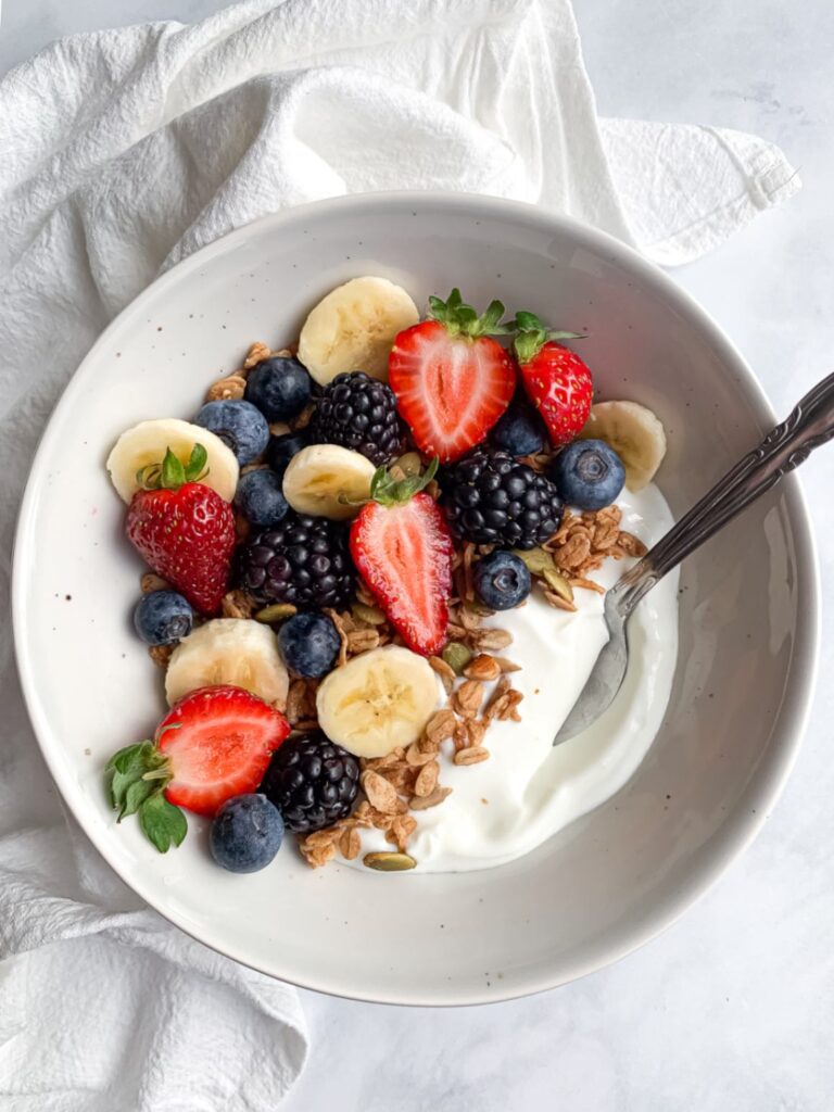 top down shot of a bowl of jurt with granola, berries and banana slices covering half of it with a spoon coming out.