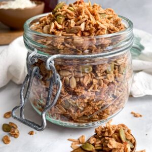 close up of a jar of maple coconut granola with shredded coconut and maple syrup in the background and a wooden spoon in the front with granola clusters on it.