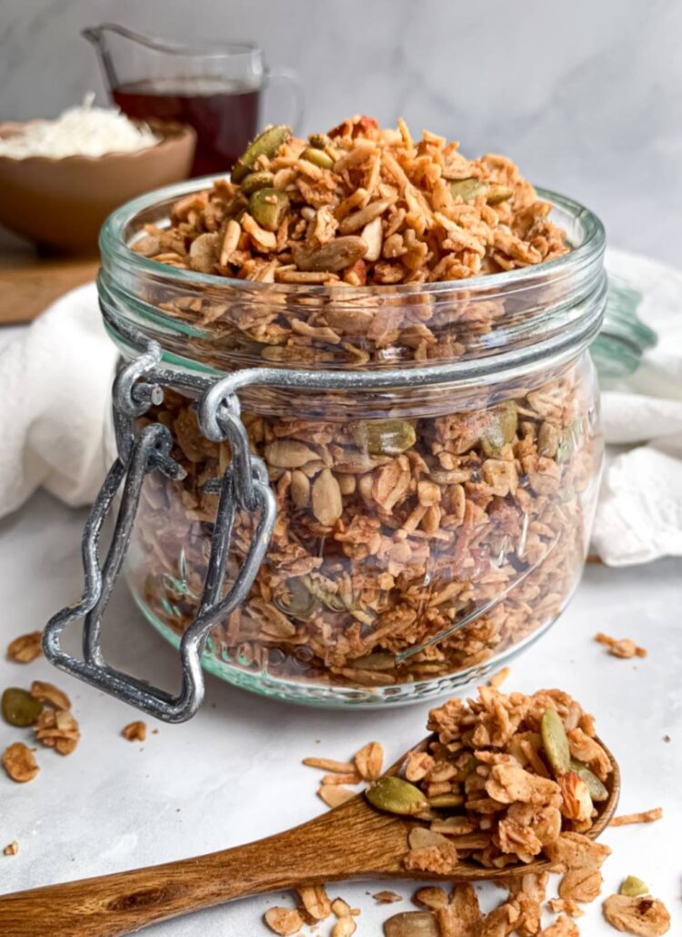 close up of a jar of maple coconut granola with shredded coconut and maple syrup in the background and a wooden spoon in the front with granola clusters on it.