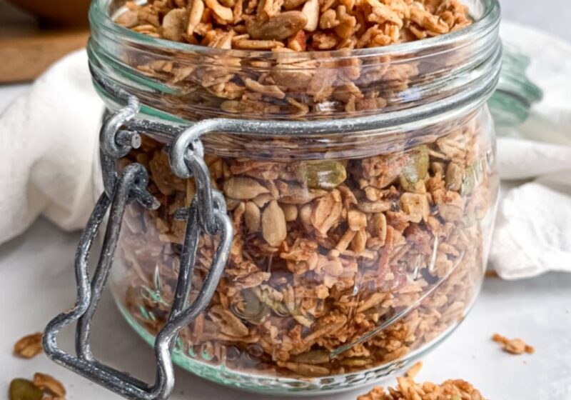close up of a jar of maple coconut granola with shredded coconut and maple syrup in the background and a wooden spoon in the front with granola clusters on it.