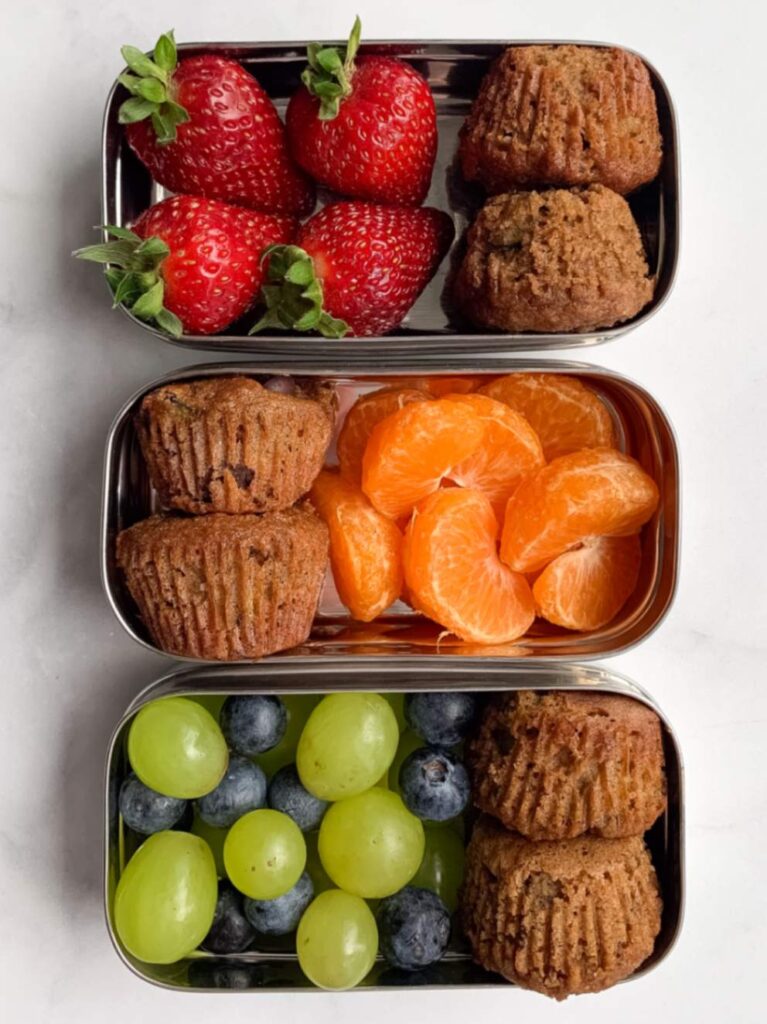 3 metal snack containers each containing two mini banana zucchini muffins and a different fruit.
