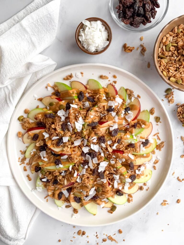 on a white plate, apple nachos topped with peanut butter, granola, seeds, raisins, and coconut flakes as a healthy snack for kids, with some ingredients in small bowls on the side.