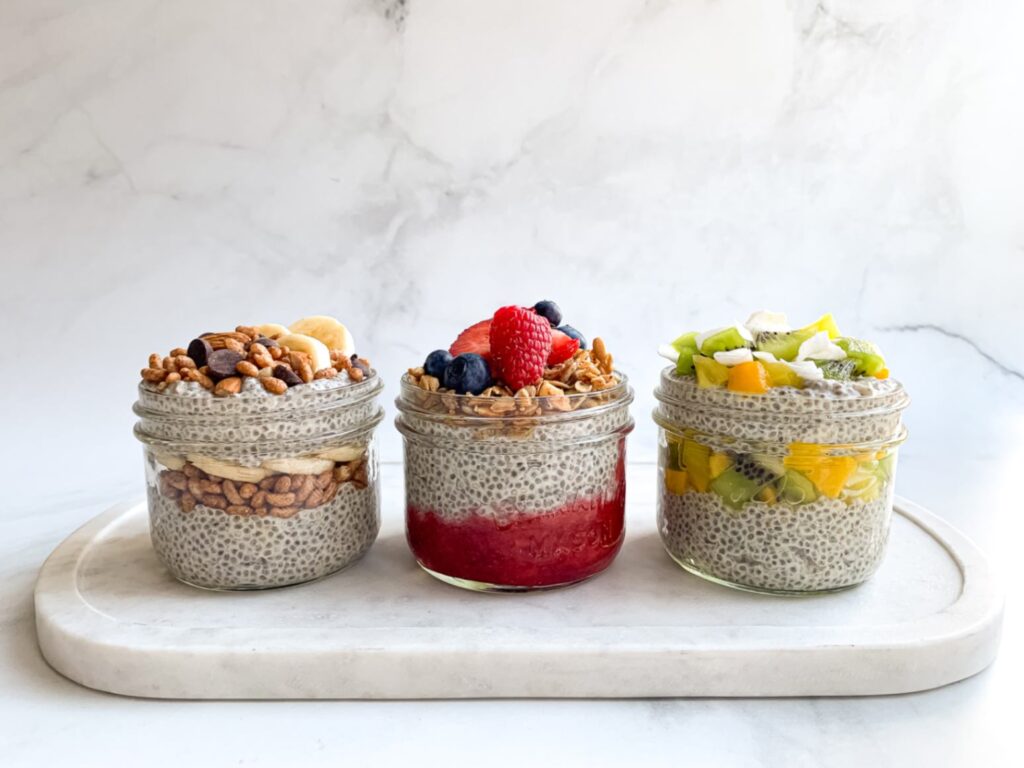 a tray with 3 jars of layered chia pudding parfaits with with different types of toppings - banana chocolate, tropical fruit and berry granola.