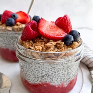 upclose of two wide-mouth half pint mason jars of chia pudding with strawberry puree on layered on the bottom and topped with granola and mixed berries on top on a marble tray.