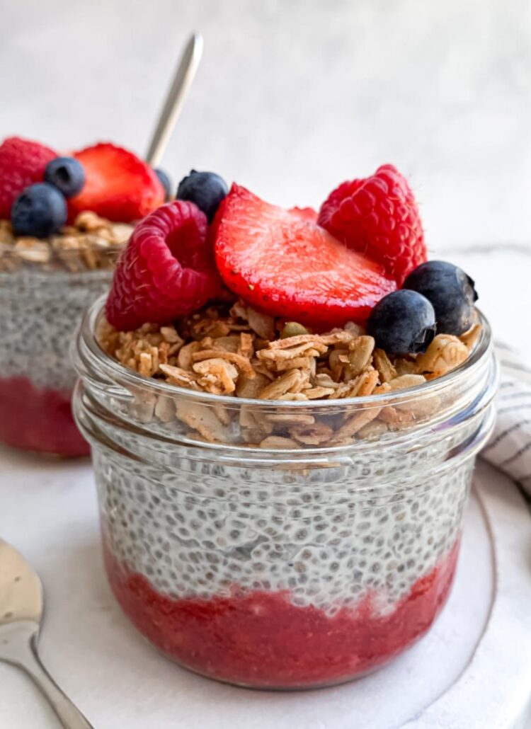 upclose of two wide-mouth half pint mason jars of chia pudding with strawberry puree on layered on the bottom and topped with granola and mixed berries on top on a marble tray.