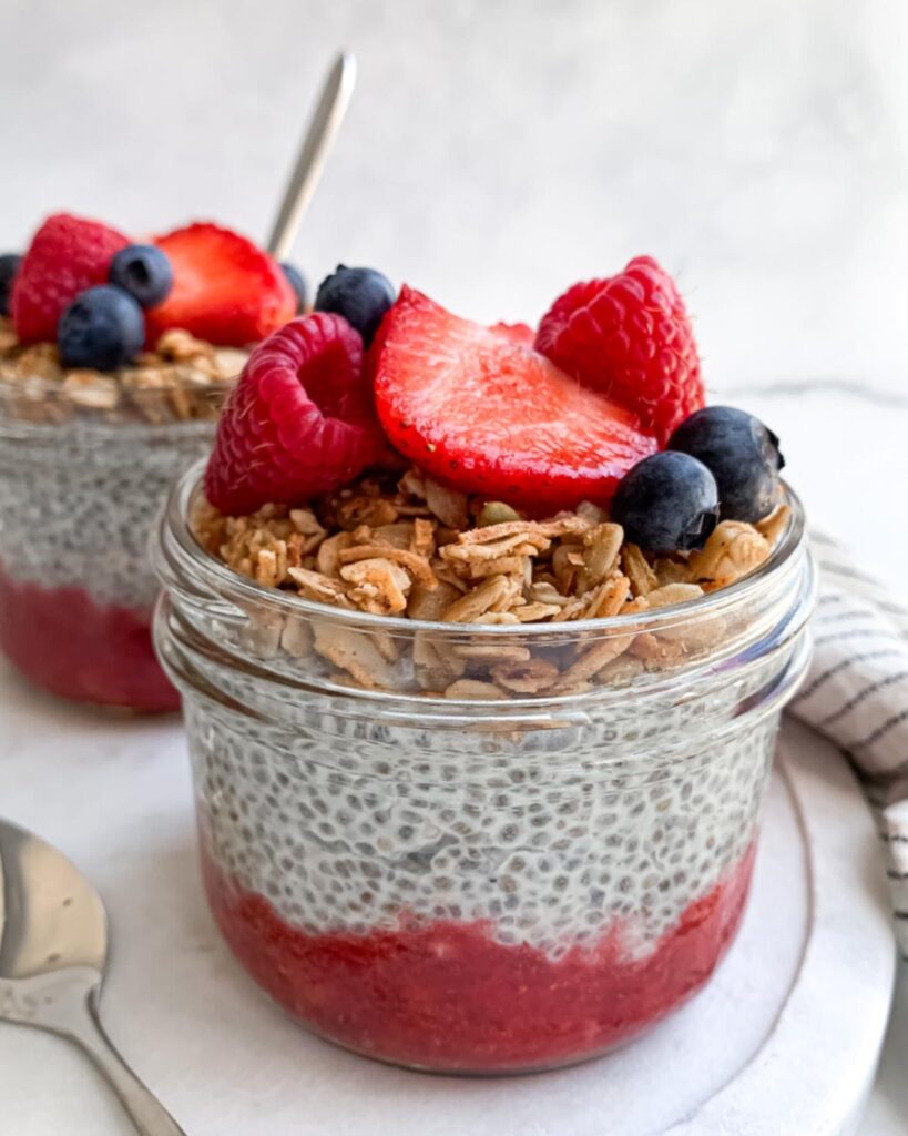 upclose of two wide-mouth half pint mason jars of chia pudding with strawberry puree on layered on the bottom and topped with granola and mixed berries on top on a marble tray.