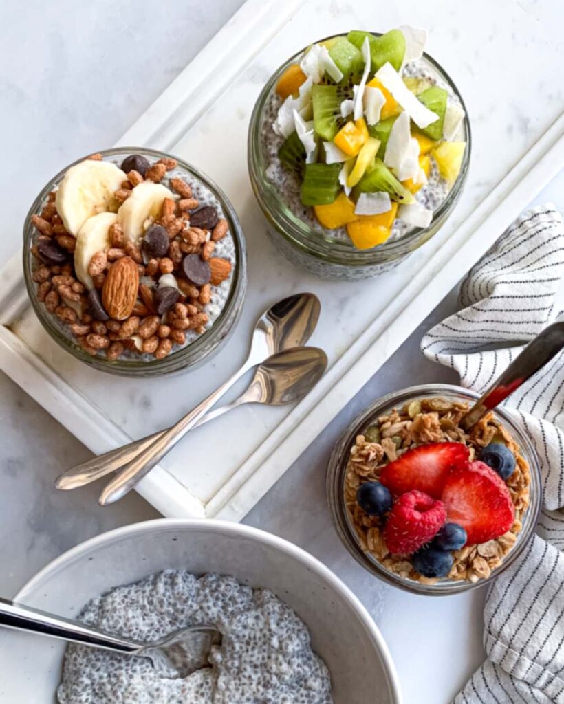 top down of 3 mason jars of chia pudding with different toppings with two on a marble tray with spoons, and a bowl of plain chia pudding with a spoon in it.