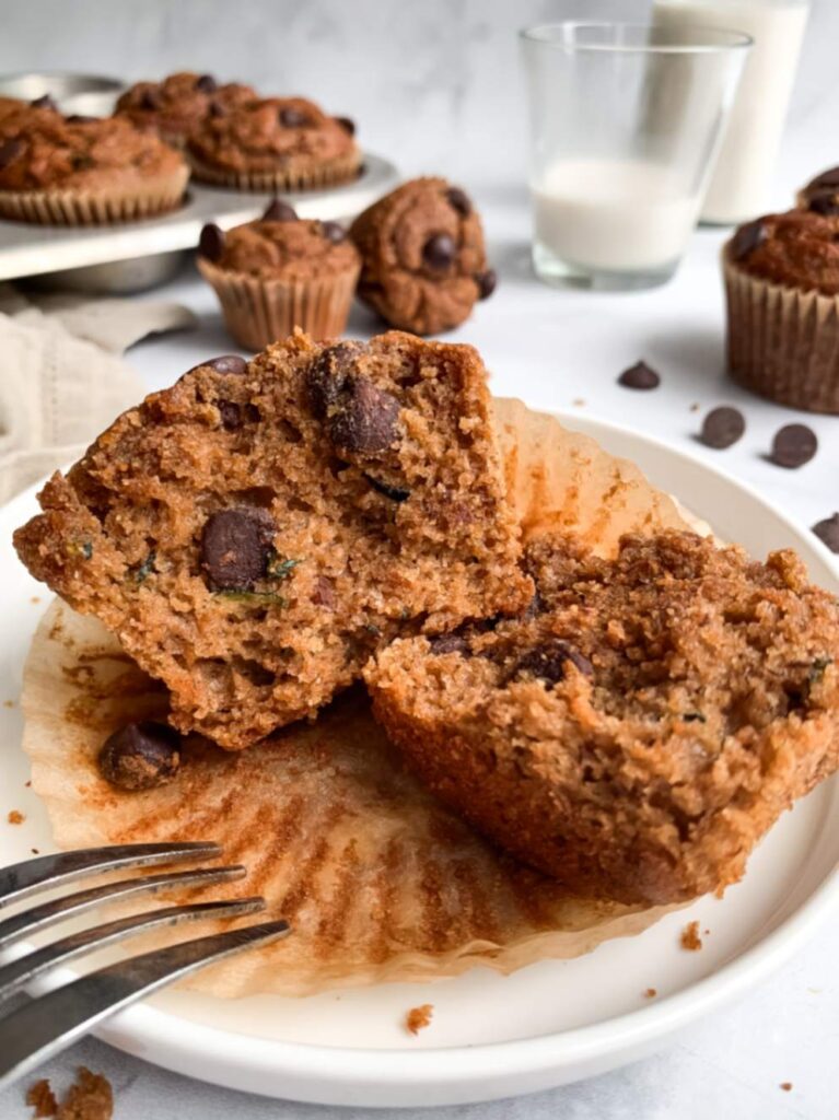 Moist gluten-free banana zucchini muffin browkn open on a plate with fork, showing the soft texture inside with glass of milk and other muffins in the background.